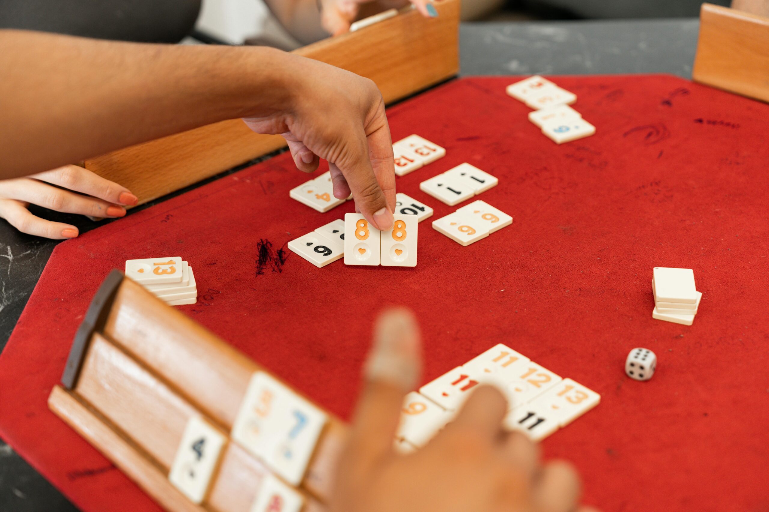 Close-up of hands playing Rummikub with numbered tiles on a red felt table.